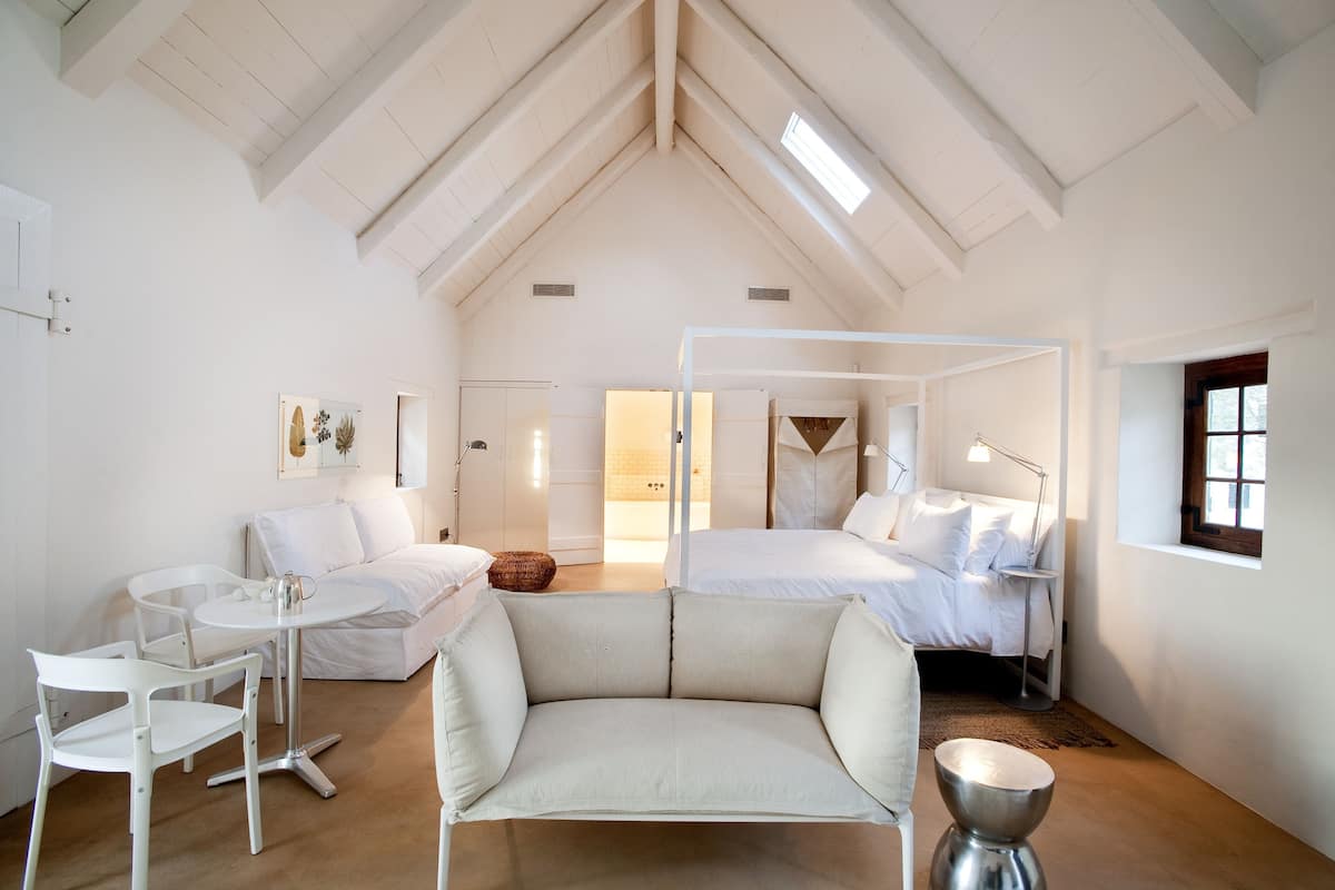 A spacious room with a high ceiling, a bed, a sofa, a small table, and a chair.