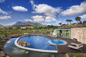 A pool with a wooden deck and lounge chairs, surrounded by rocks and plants, with mountains in the background. - Babylonstoren (Franschhoek)
