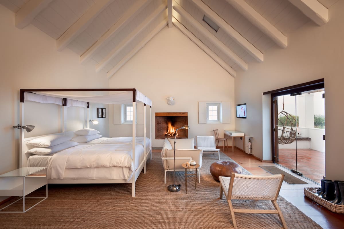 A bedroom with a four-poster bed, a fireplace, a balcony with a chair, and a view of the outdoors.