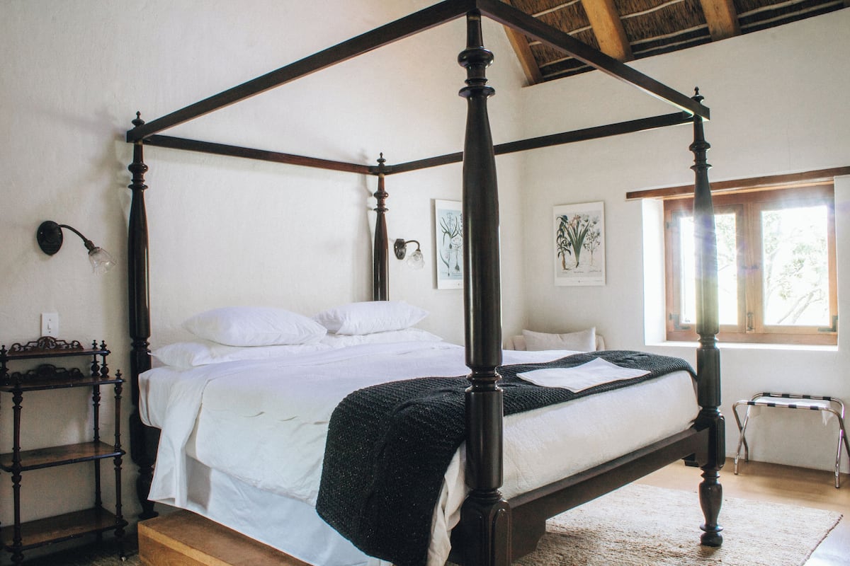 A four-poster bed with a wooden frame, white bedding, and a black throw blanket. There is a small bedside table with a lamp, a framed botanical print on the wall, and a window with natural light.
