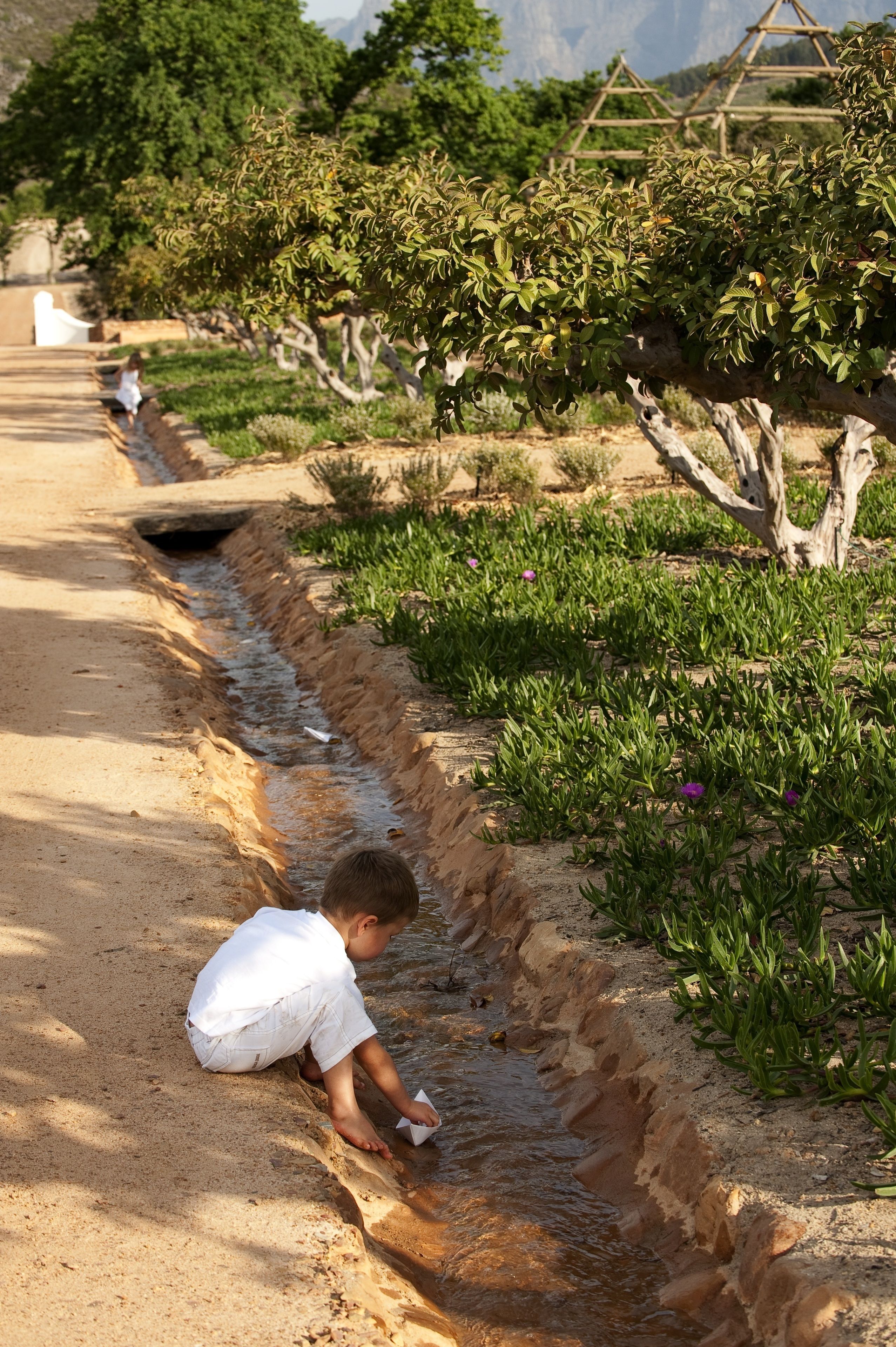 A child playing by a water channel in a garden with trees and a wooden structure in the background.