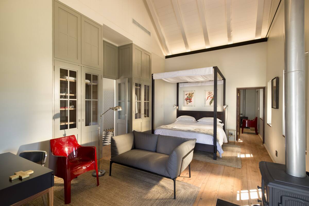 A modern bedroom with a four-poster bed, a red chair, a sofa, and a wooden desk.