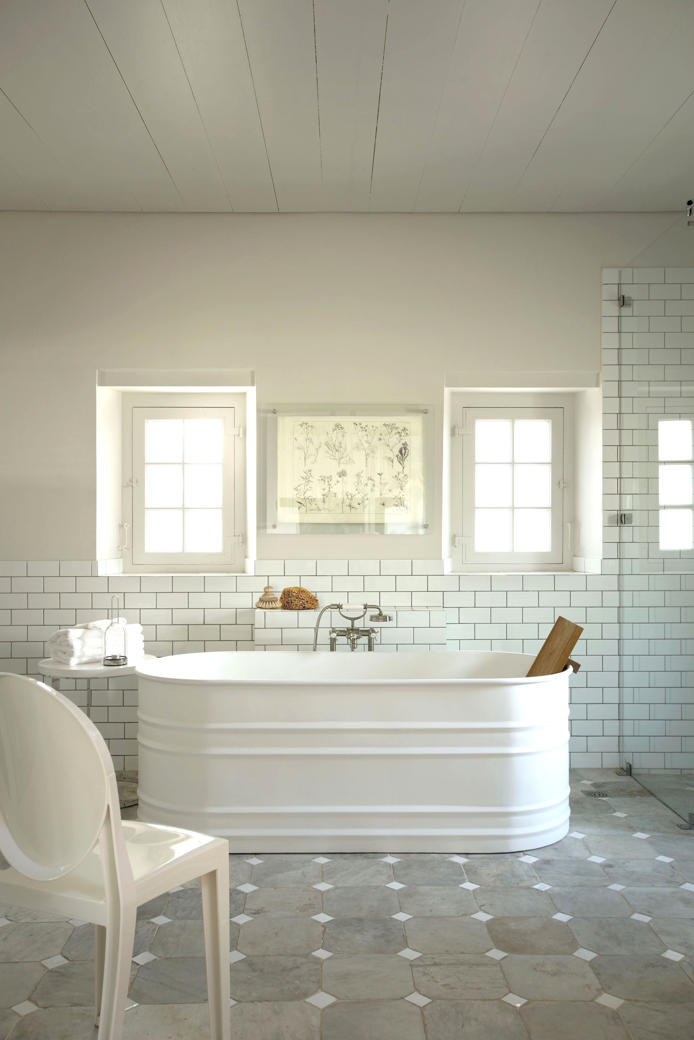A modern bathroom with a freestanding bathtub, two windows, a white chair, and a tiled floor.