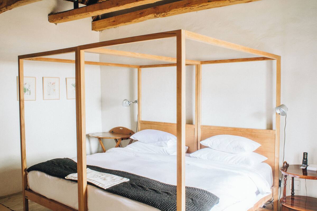 A four-poster bed with white linens, a bedside table with a lamp, and framed pictures on the wall.
