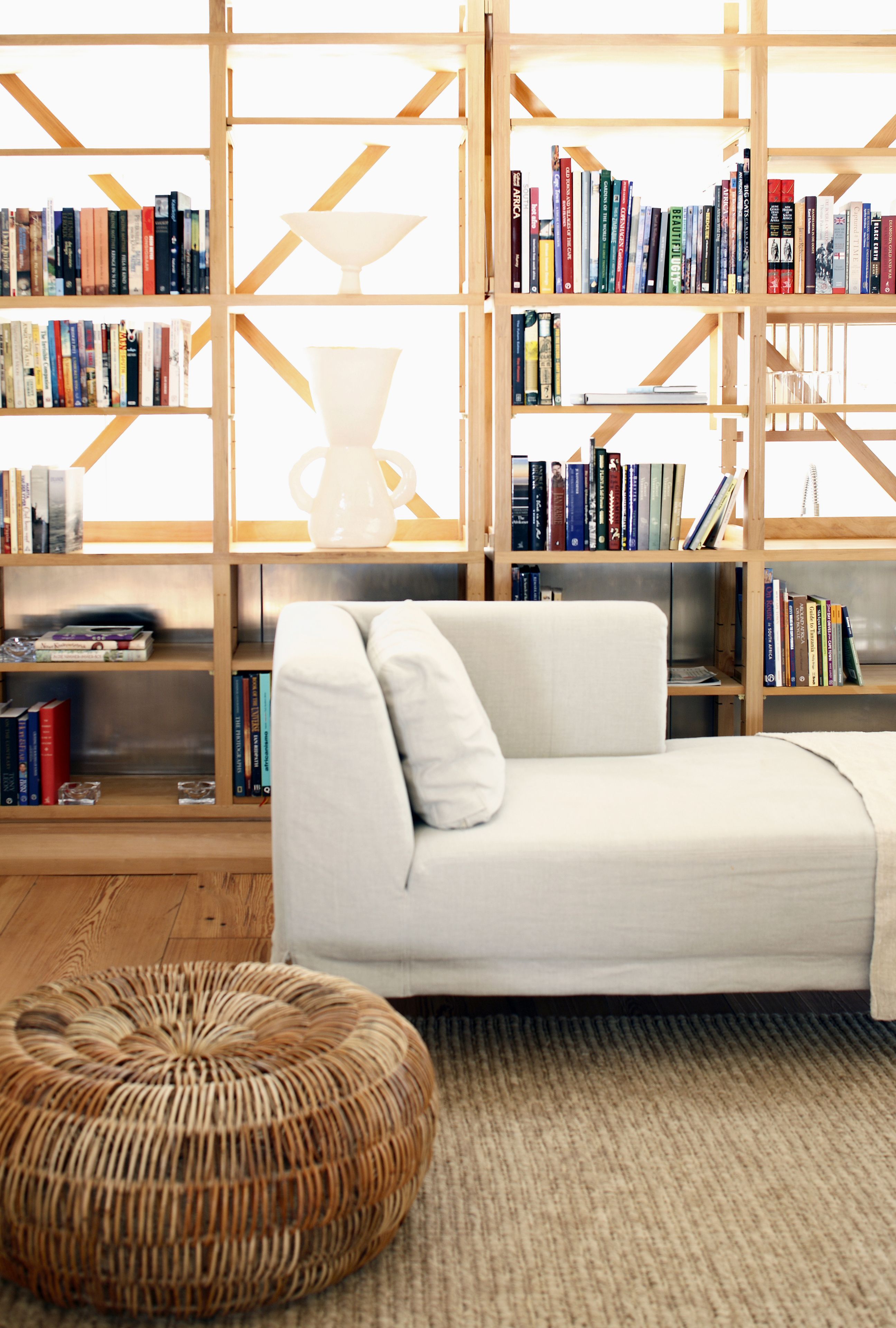 Una habitación con un sofá blanco, un otoman de mimbre y estanterías de madera llenas de libros.