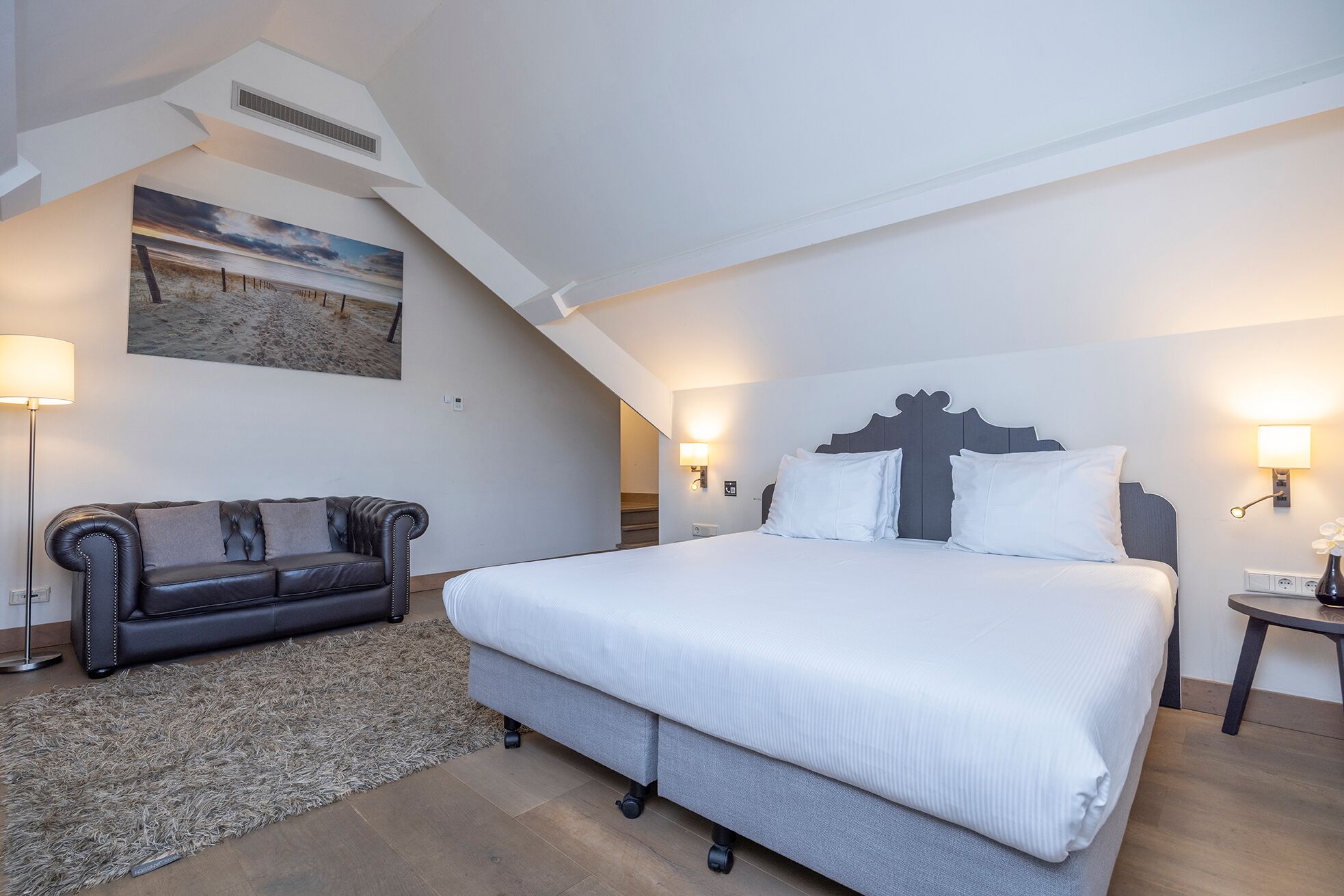 A bedroom with a large bed, a dark leather sofa, a painting, and a staircase.