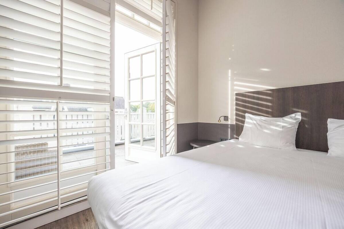 A hotel room with a large bed, a nightstand, a window with blinds, and a view of the outside.
