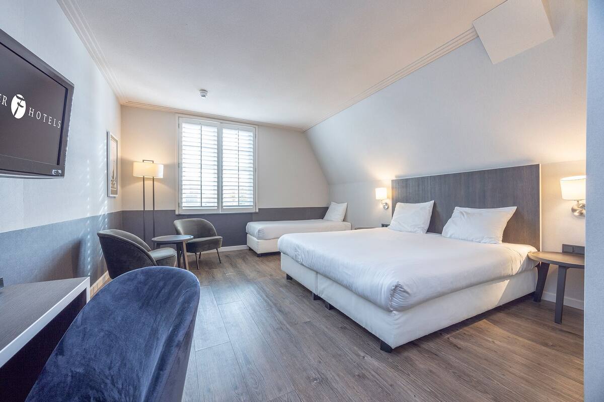 A hotel room with a large bed, two bedside tables, a desk, a chair, and a window with blinds.