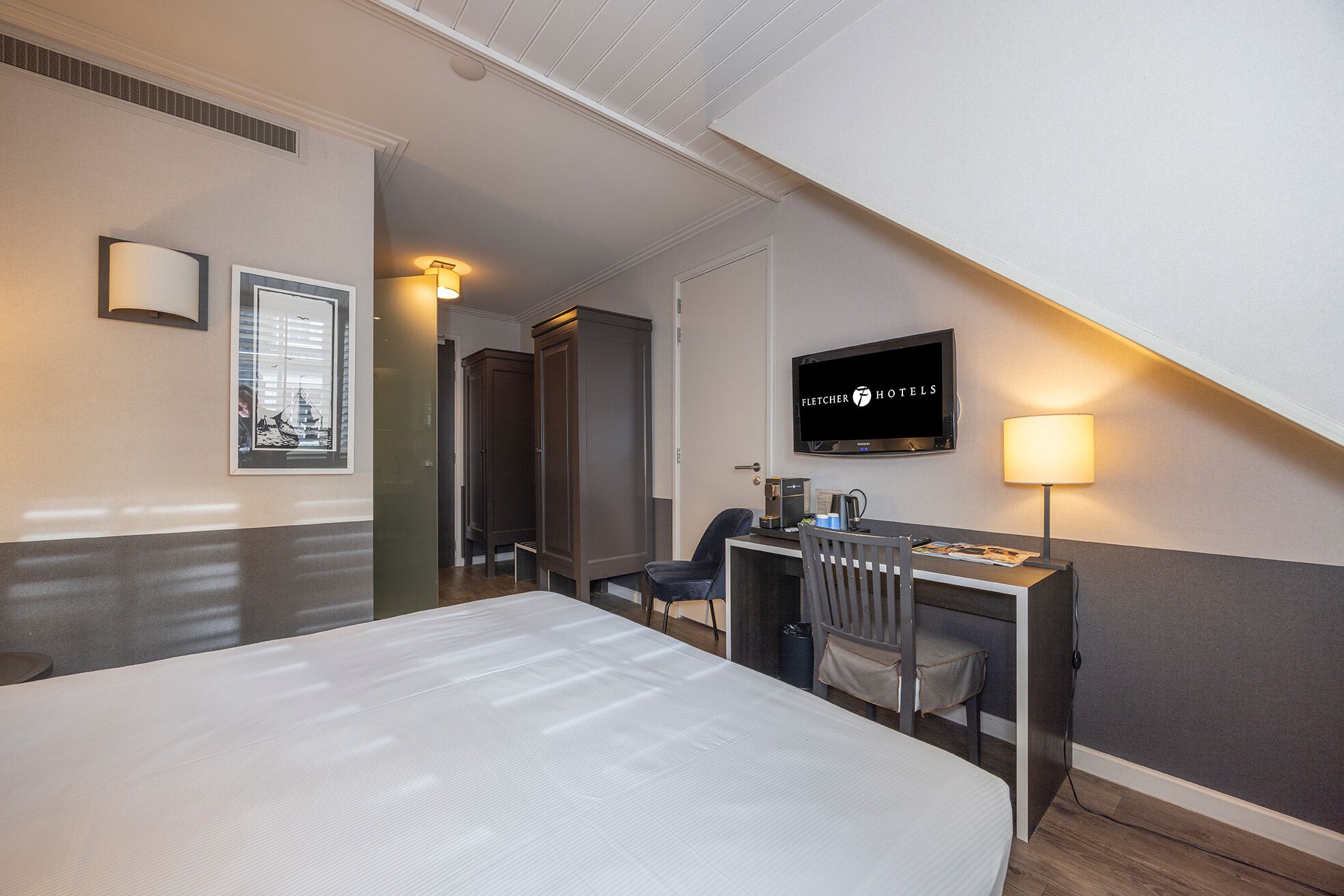 Une chambre d’hôtel équipée d’un lit, d’un bureau, d’une chaise et d’un téléviseur affichant le logo « Fletcher Hotels ».