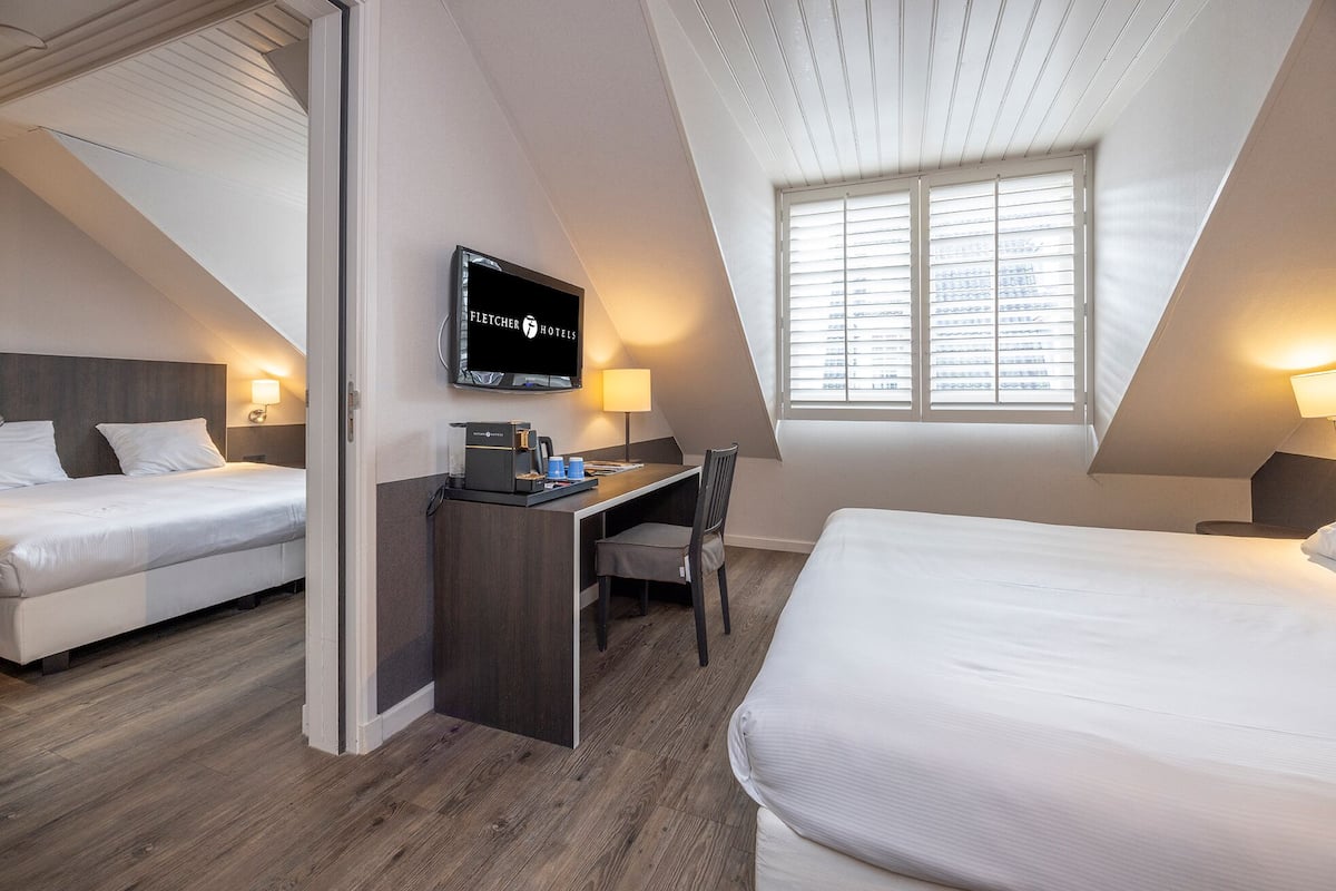A hotel room with two beds, a desk, a TV, and a window with blinds.