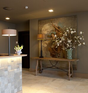 A reception area with a reception desk, a lamp, a painting, and a vase with flowers. - Fletcher Nautisch Kwartier (Huizen)