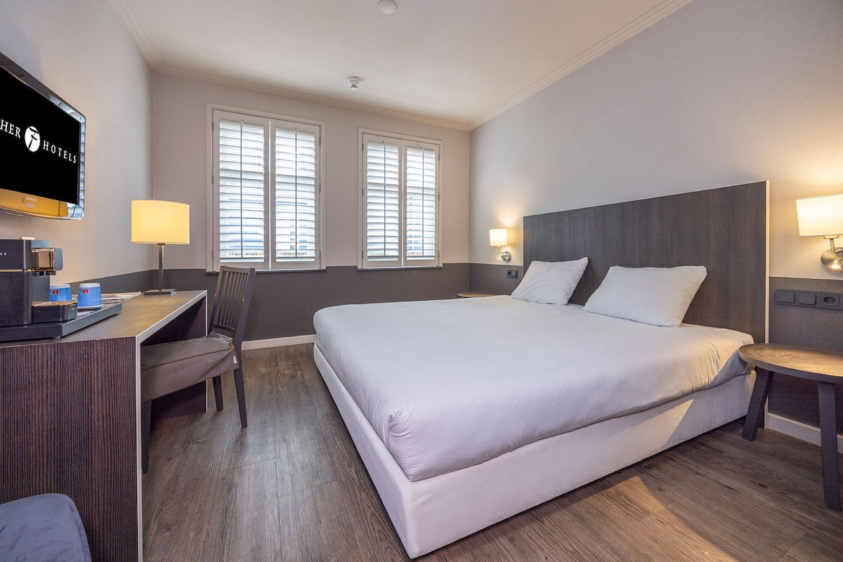 A hotel room with a large bed, a desk with a coffee machine, two bedside lamps, and a TV mounted on the wall.