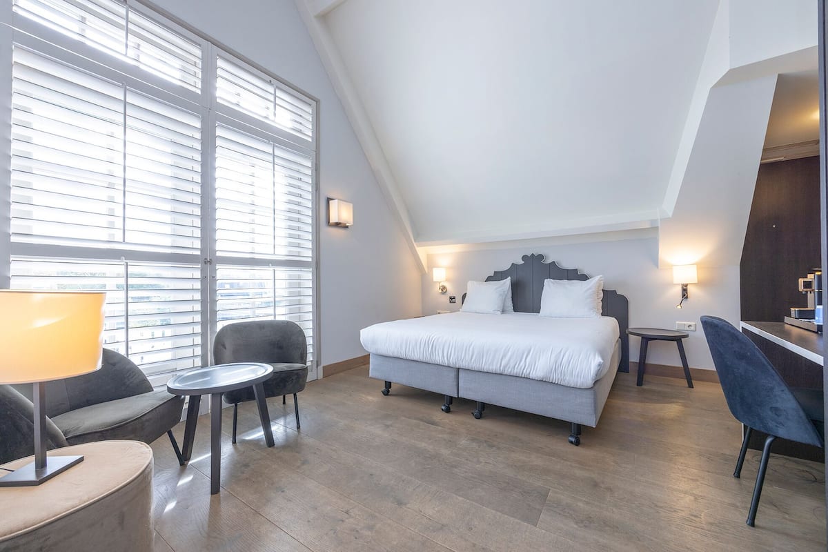 A modern hotel room with a large bed, a desk, a chair, and a window with blinds.