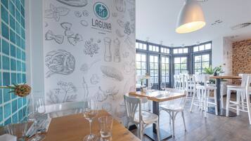 Een restaurantinterieur met een houten tafel, witte stoelen en een muur versierd met afbeeldingen van eten.