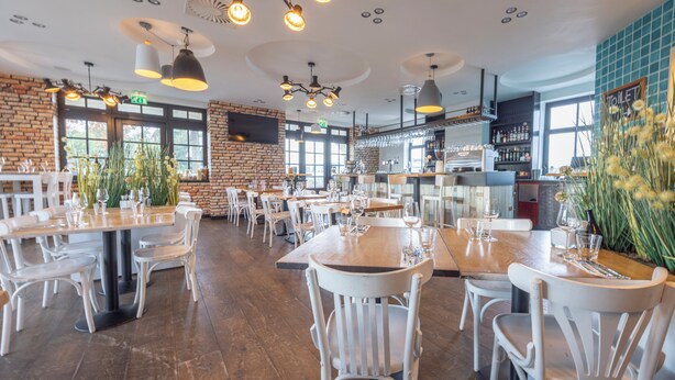 A spacious restaurant with high ceilings, exposed brick walls, and modern pendant lighting. Tables are set with white chairs and wine glasses, and there are large windows allowing natural light. - Fletcher Nautisch Kwartier (Huizen)