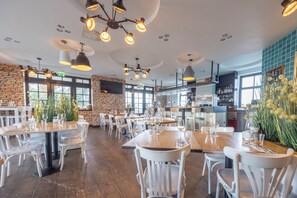 Un restaurante espacioso con techos altos, paredes de ladrillo visto e iluminación colgante moderna. Las mesas están dispuestas con sillas blancas y copas de vino, y hay grandes ventanales que permiten la entrada de luz natural.