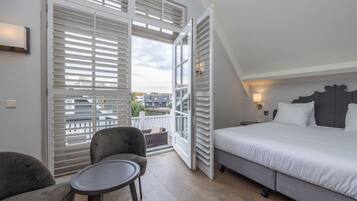 Un dormitorio con cama, una silla, una mesita y un balcón con contraventanas.