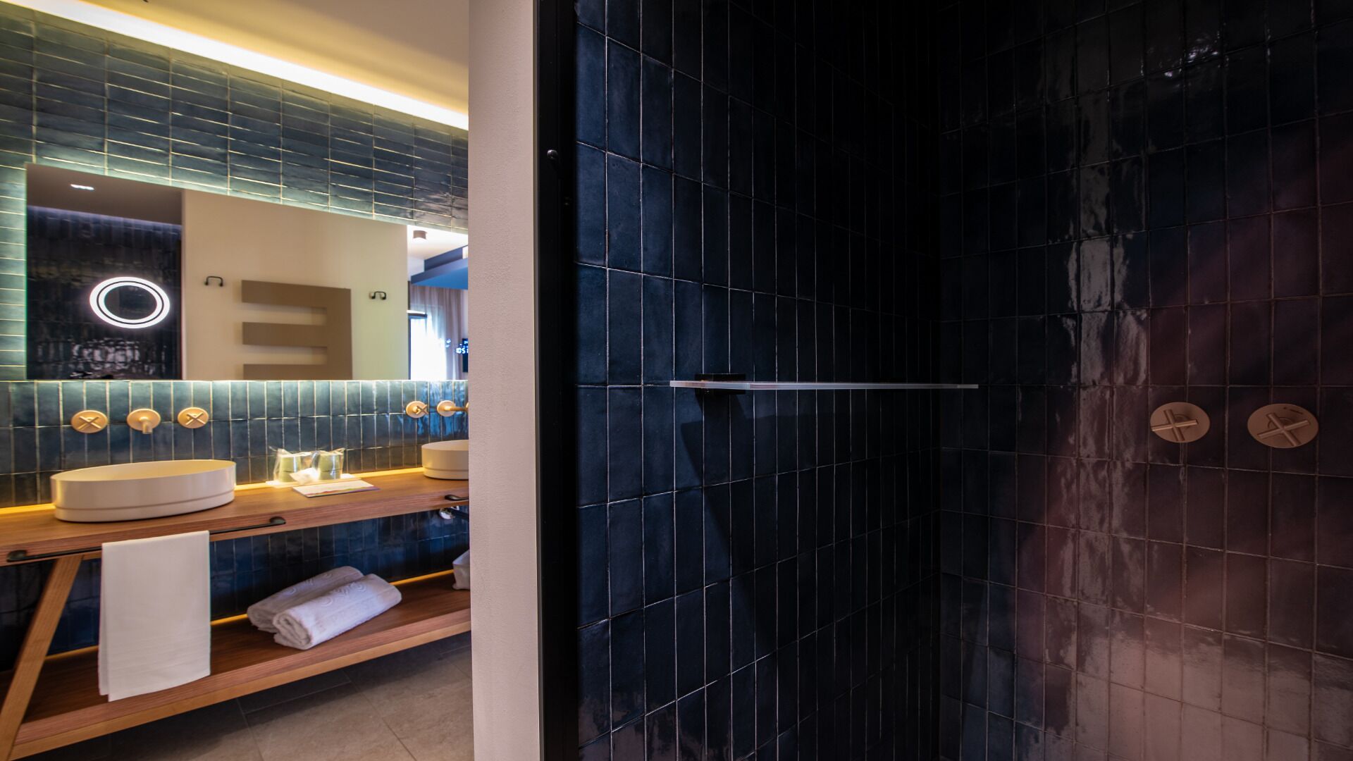 Une salle de bain moderne avec une douche aux carreaux foncés, un meuble-lavabo doté d’un miroir rond et une étagère en bois où sont posées des serviettes.