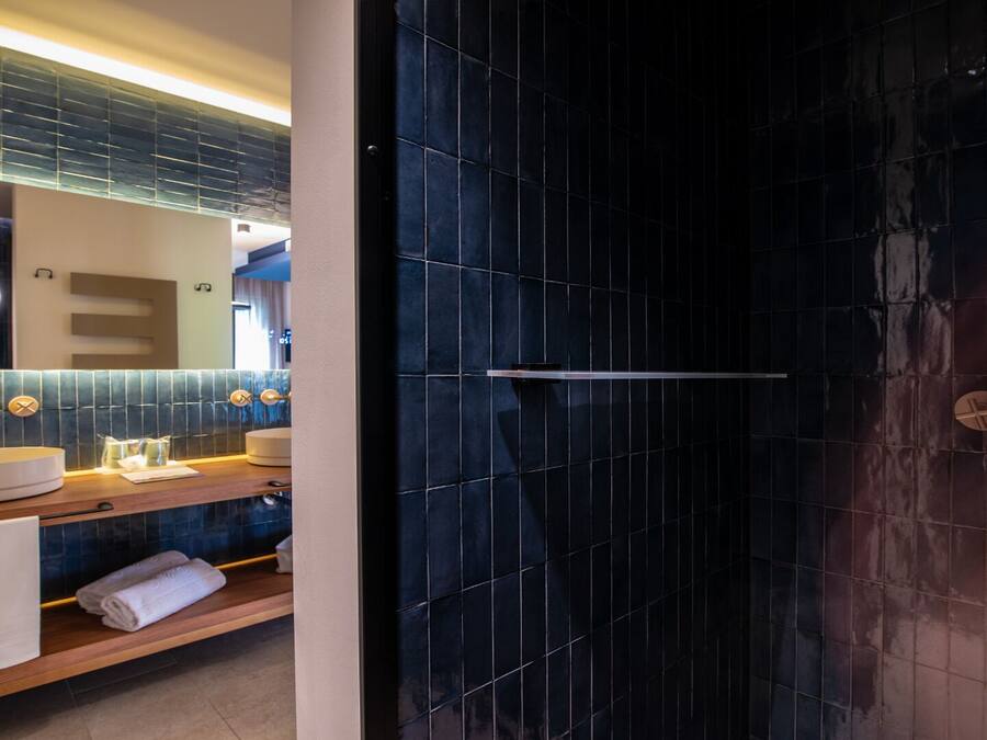 Une salle de bain moderne avec une douche aux carreaux foncés, un meuble-lavabo doté d’un miroir rond et une étagère en bois où sont posées des serviettes.