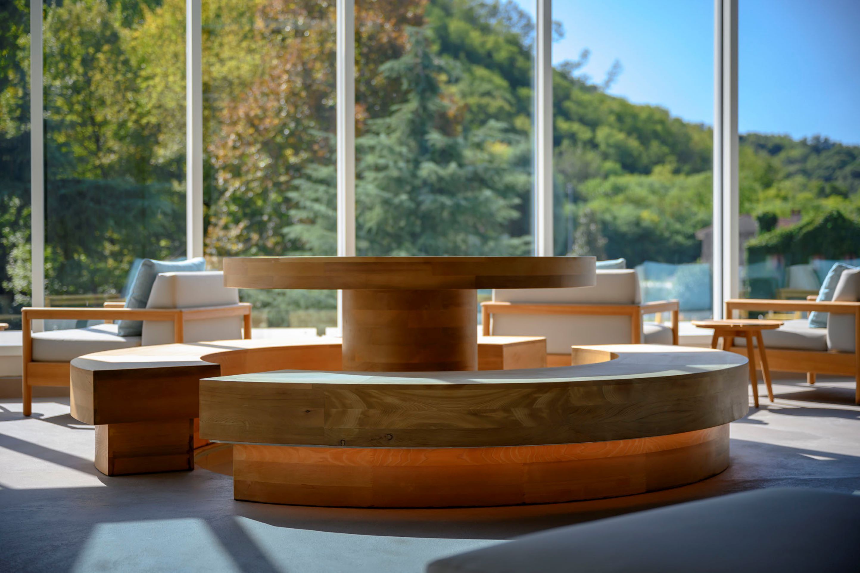 A modern outdoor seating area with a central wooden table and multiple chairs, surrounded by large windows offering a view of a forested hillside.