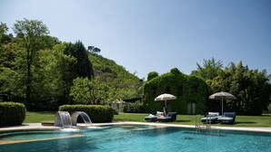 Piscina con sillas de descanso y sombrillas, rodeada de vegetación y una colina.