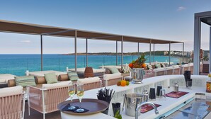 Un espace extérieur aménagé avec des sièges, baigné de lumière, offrant une vue sur la mer, comprenant des fauteuils confortables, un bar avec un plateau en verre et un grand pichet en métal.
