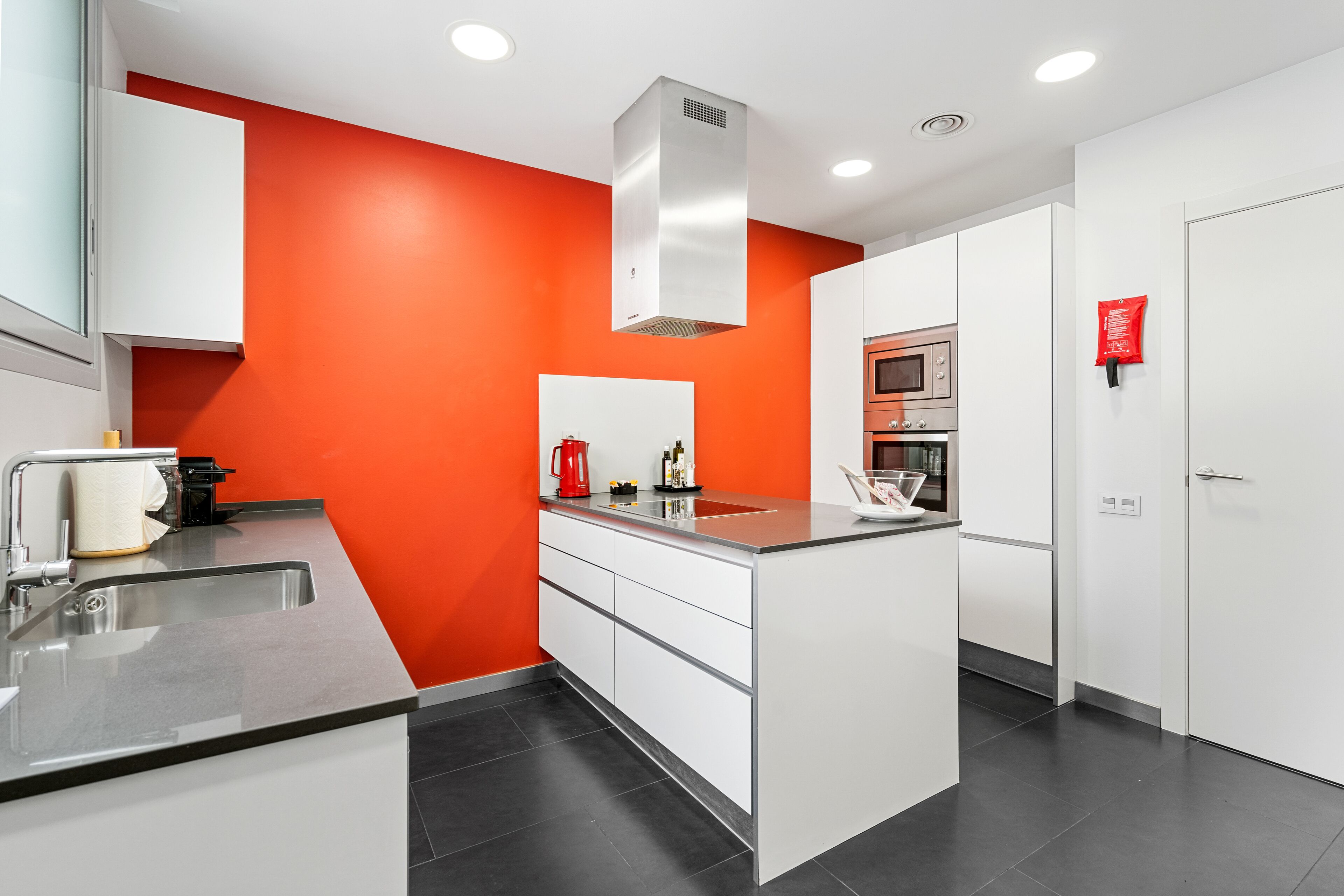 A modern kitchen with white cabinets, a red accent wall, and a stainless steel sink.