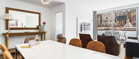 A modern hotel room with a large dining table, comfortable seating, a large mirror, and a view of a historic building through the window.