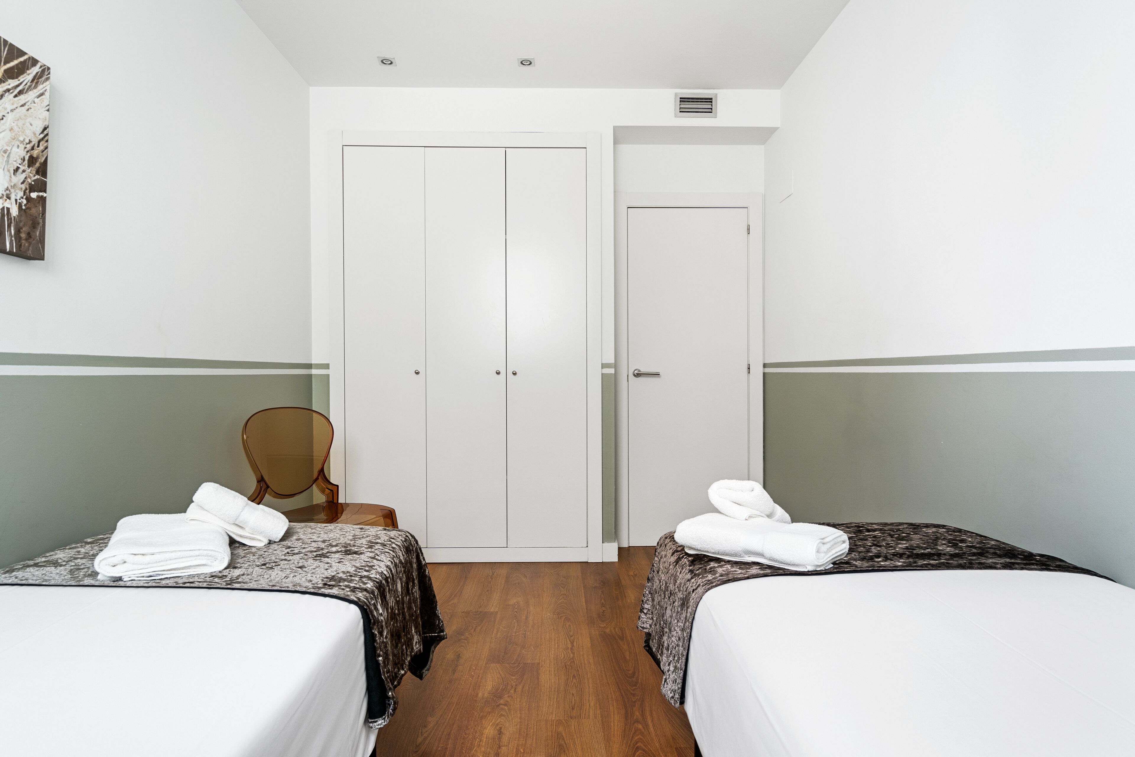 A hotel room with two beds, a wooden chair, a white door, and a painting on the wall.