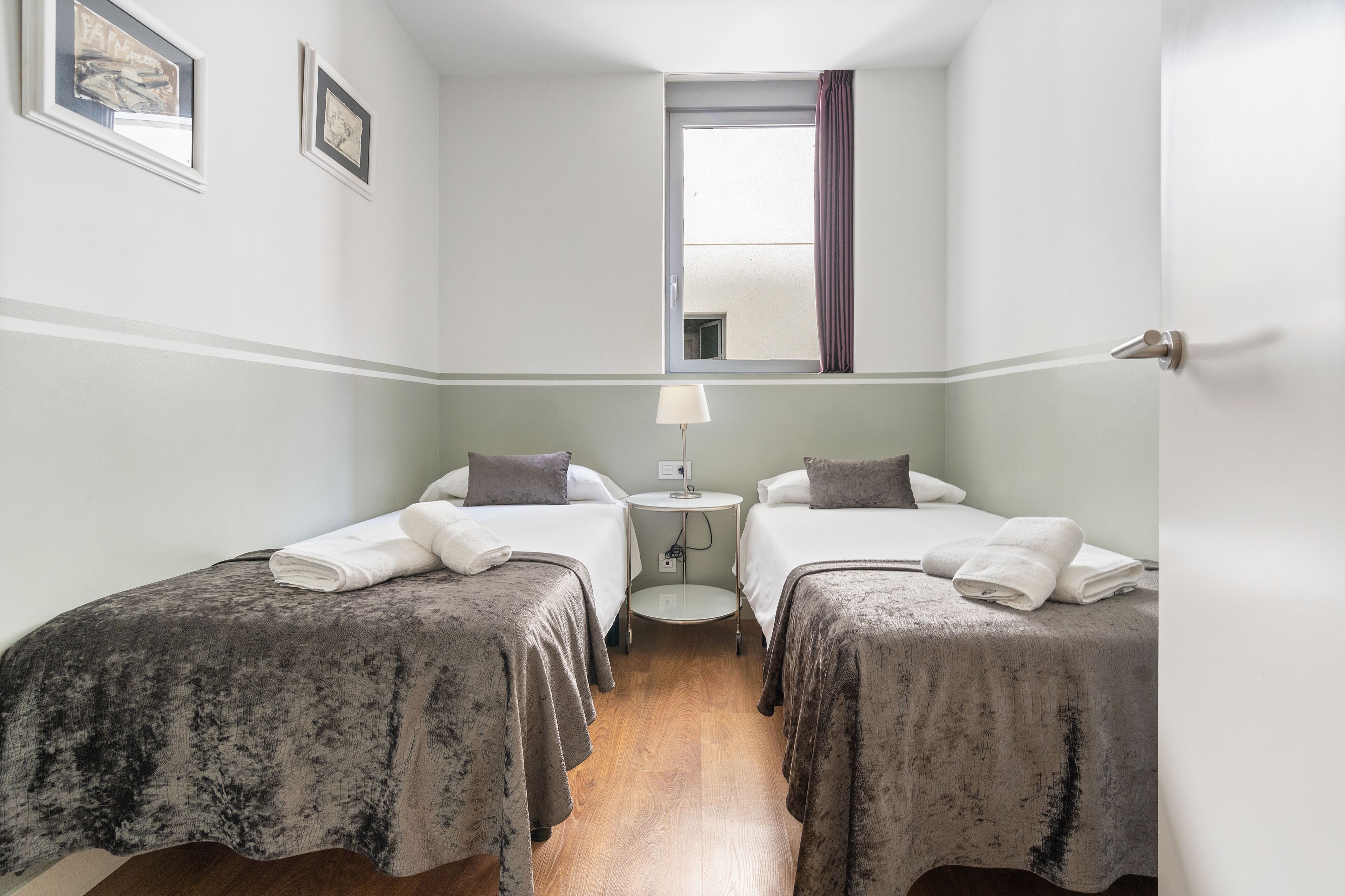 A hotel room with two beds, a small round table with a lamp, a window with curtains, and artwork on the walls.