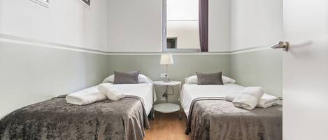A hotel room with two beds, a small round table with a lamp, a window with curtains, and artwork on the walls.