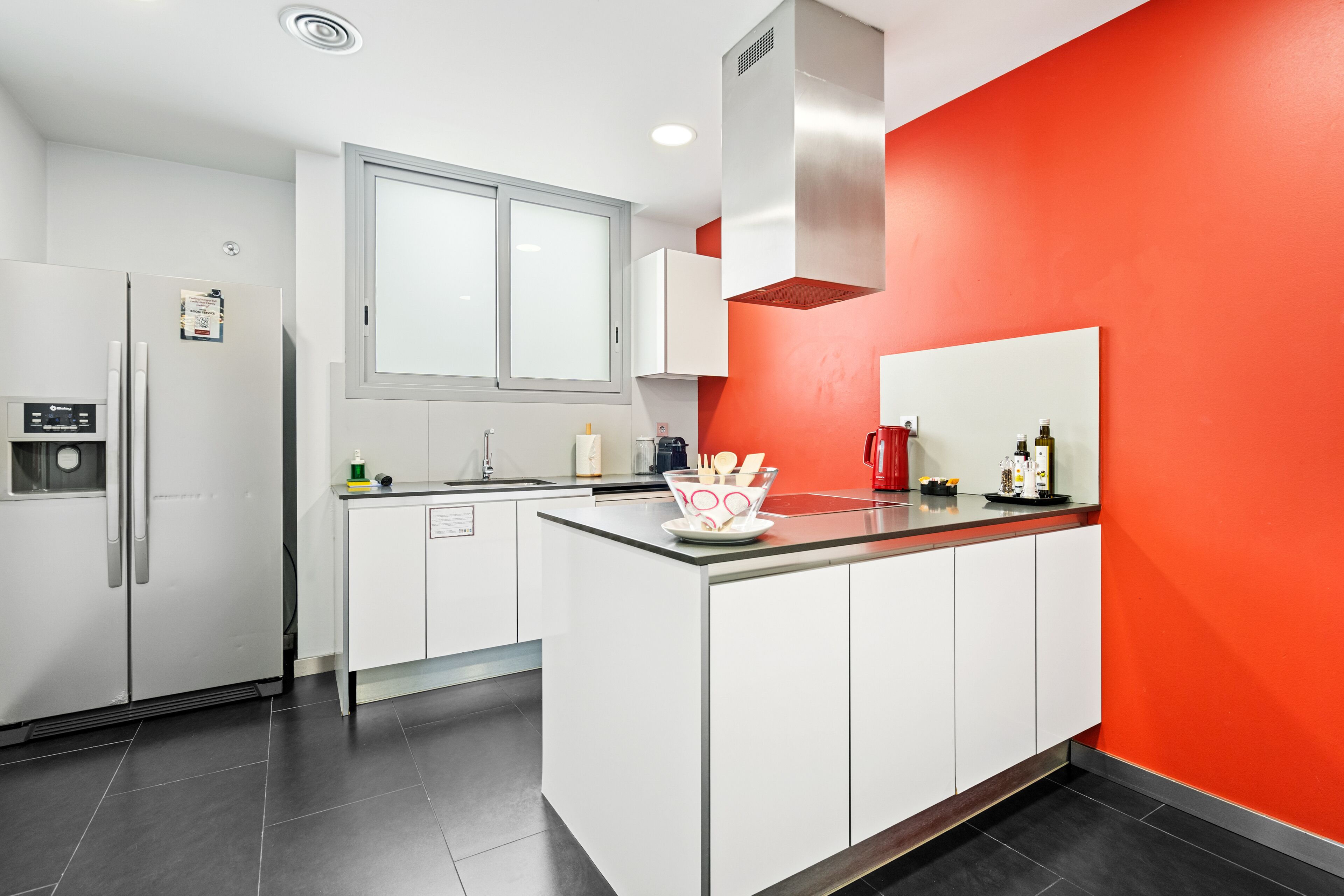 A modern kitchen with white cabinets, a red accent wall, and a stainless steel refrigerator.