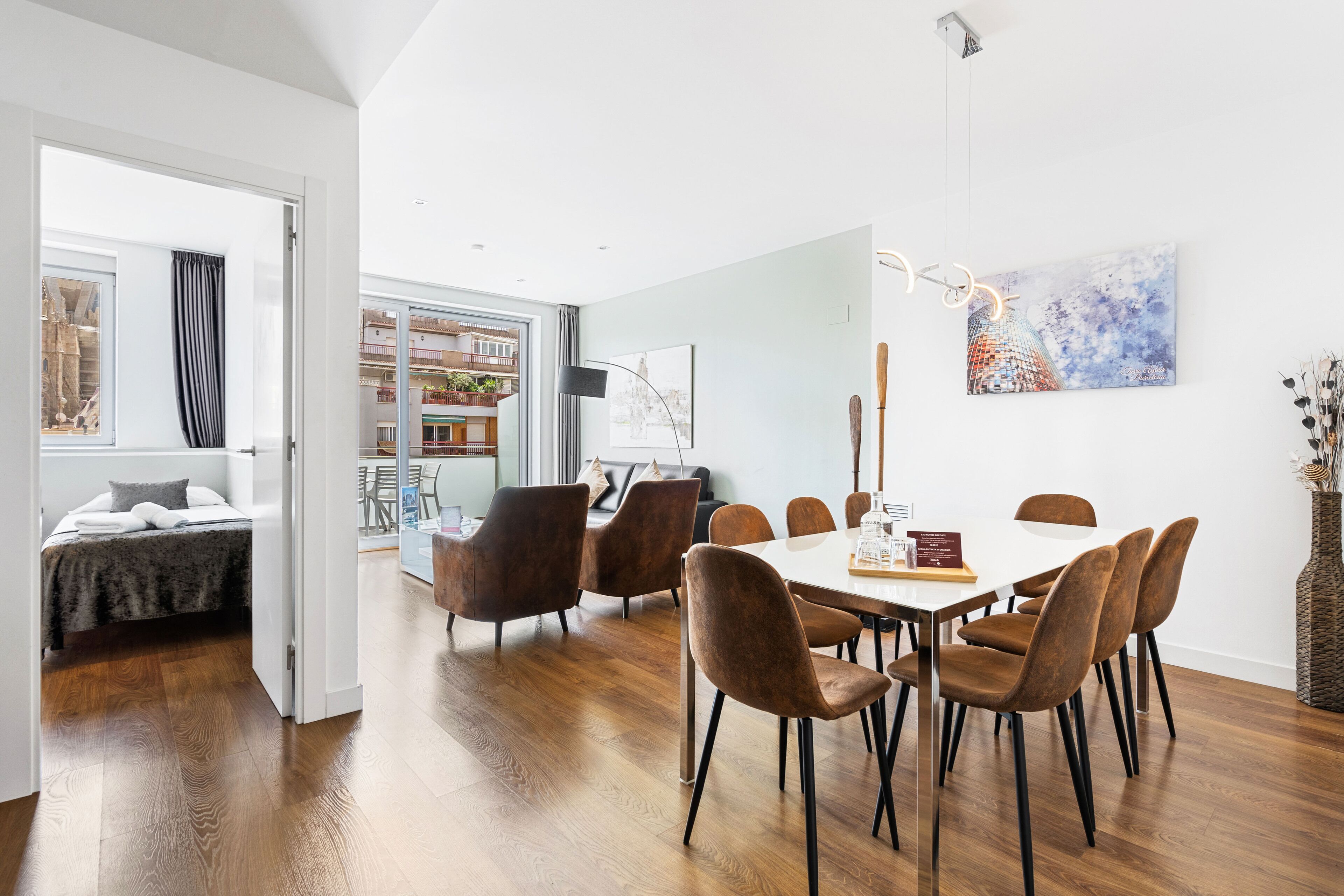 Een moderne eetruimte met een witte tafel en bruine stoelen, een schilderij aan de muur en uitzicht op een balkon met planten.