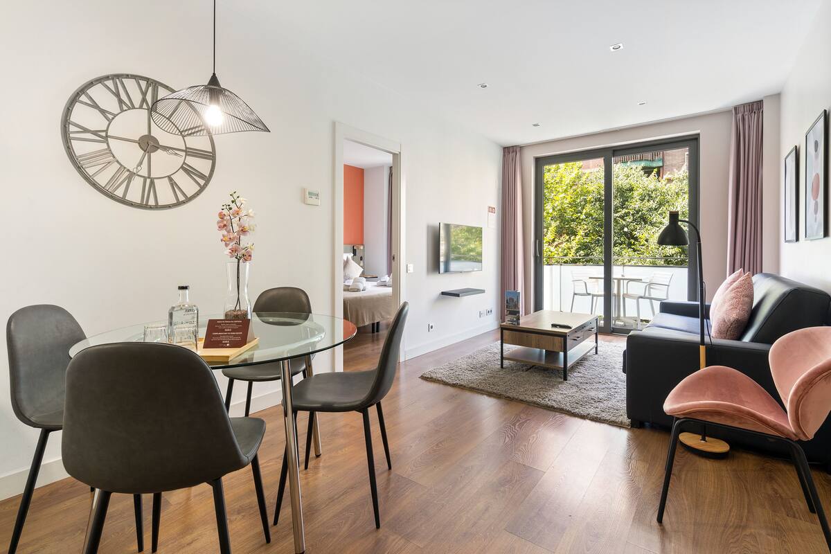 Un salon moderne avec une table à manger en verre, des chaises noires et roses, un canapé noir, une grande horloge murale et un téléviseur.