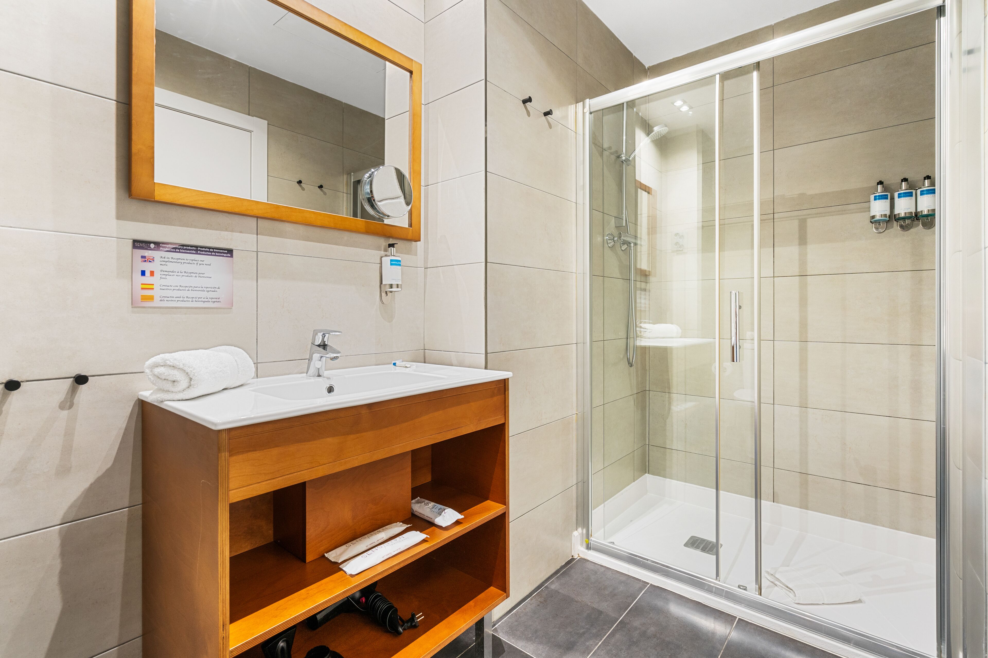 Un baño moderno con tocador de madera, cabina de ducha con mampara de vidrio y espejo con marco de madera.