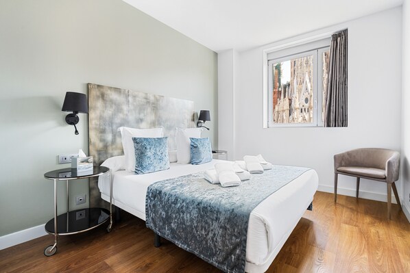 A modern hotel room with a large bed, a bedside table with a lamp, a chair, and a window with a city view. - Sensation Sagrada Familia (Barcelona)