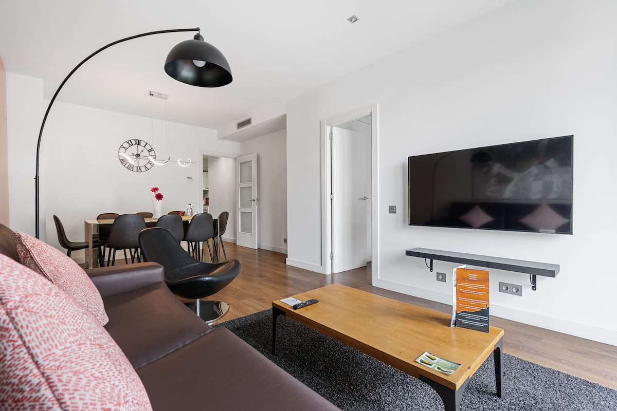 Un salon moderne avec un canapé, une table basse, un téléviseur fixé au mur et, au fond, un coin repas.