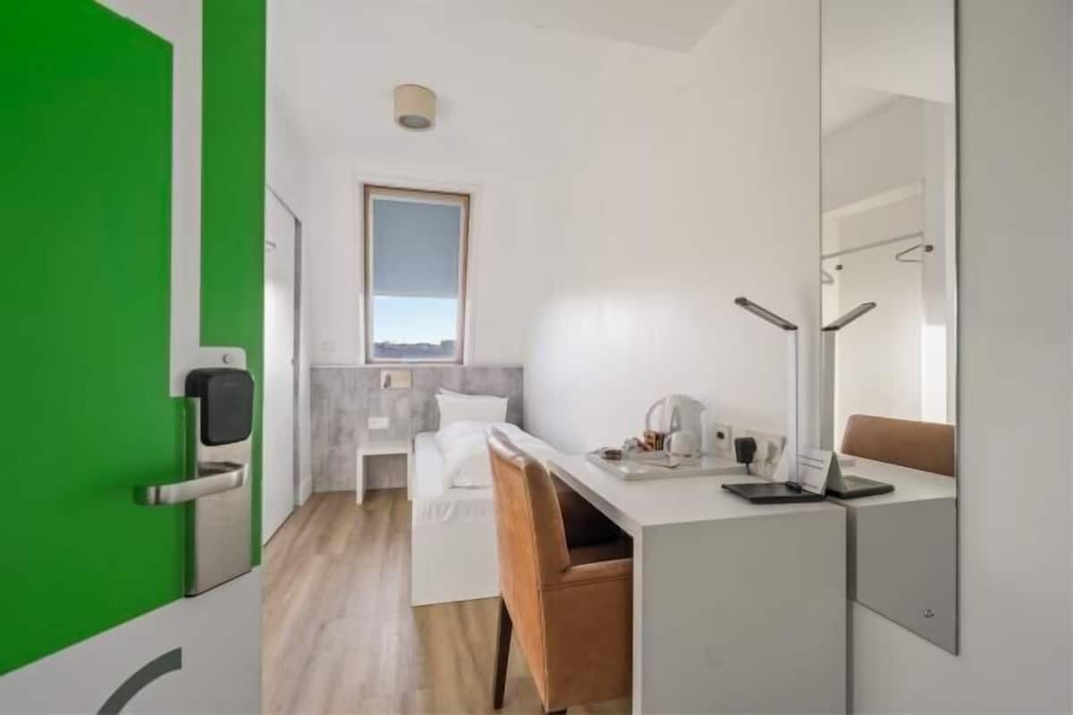 Et moderne hotelværelse med en grøn dør, en seng med hvide sengetøj, et skrivebord med en bærbar computer, en stol og et vindue med udsigt.
