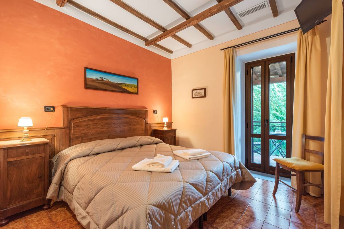Un dormitorio con una cama grande, cabecera de madera, dos mesitas de noche con lámparas, un cuadro en la pared y vistas a través de un ventanal.