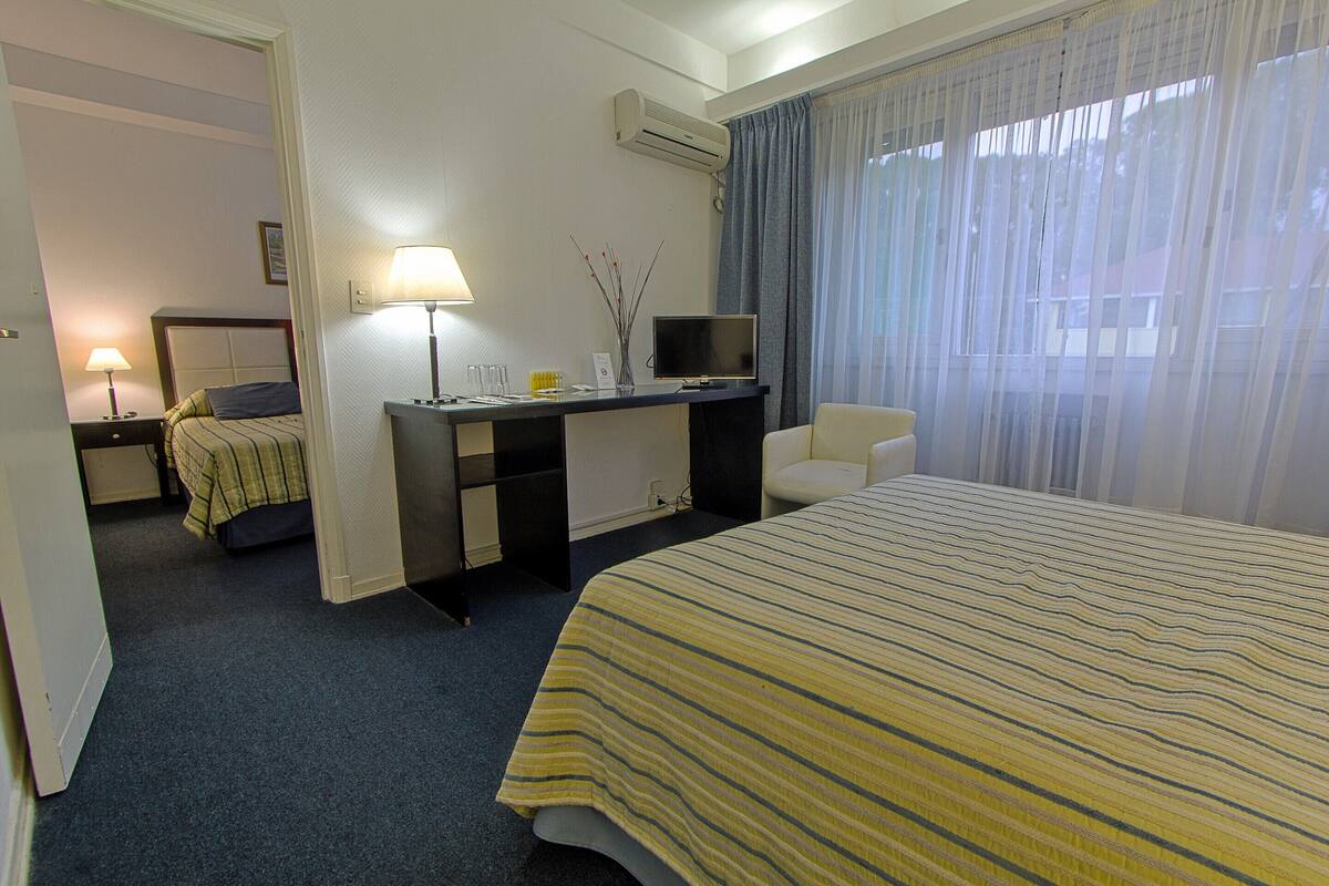 A hotel room with a bed, a desk with a computer, a chair, a lamp, and a window with curtains.