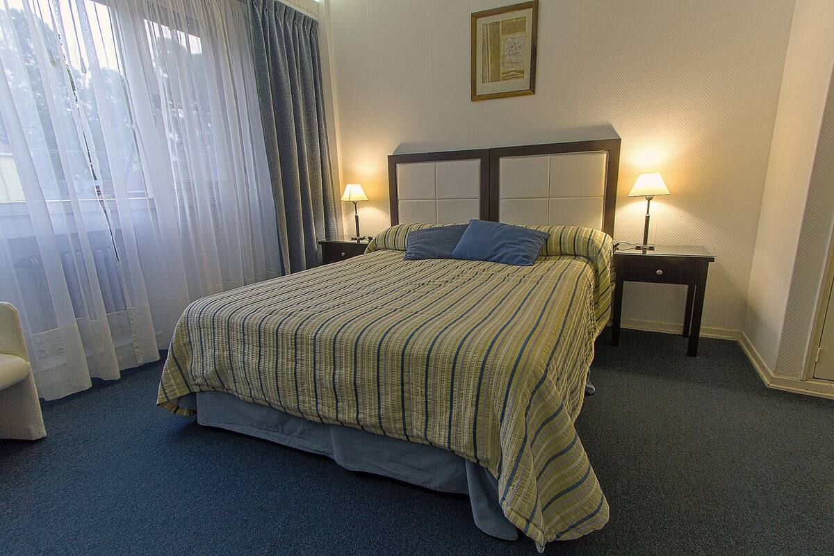 A hotel room with a bed, two bedside tables with lamps, a chair, and a window with curtains.