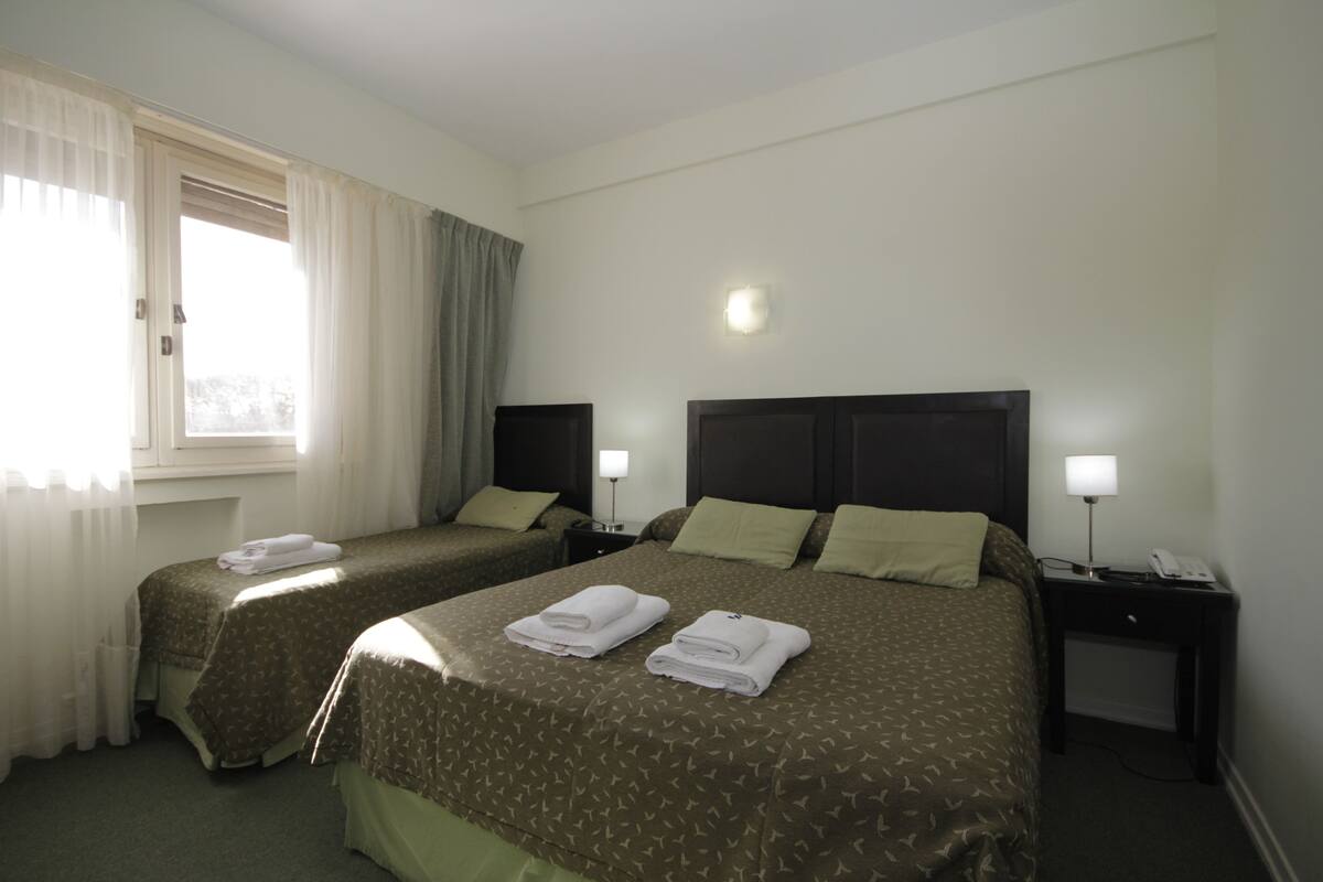 A hotel room with two beds, a nightstand with a lamp, and a window with curtains.