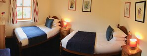Two single beds with wooden frames, white bedding, and dark blue throw blankets. Each bed has a bedside table with a red lamp. The room has a window with curtains, and there are framed artworks on the wall. - Dolphin Bay Suites (Inverness)