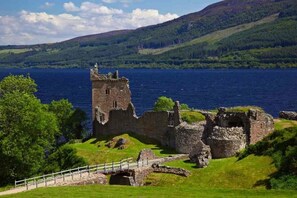 A historic castle ruin with a stone bridge leading to it, surrounded by lush green hills and a large lake. - Dolphin Bay Suites (Inverness)