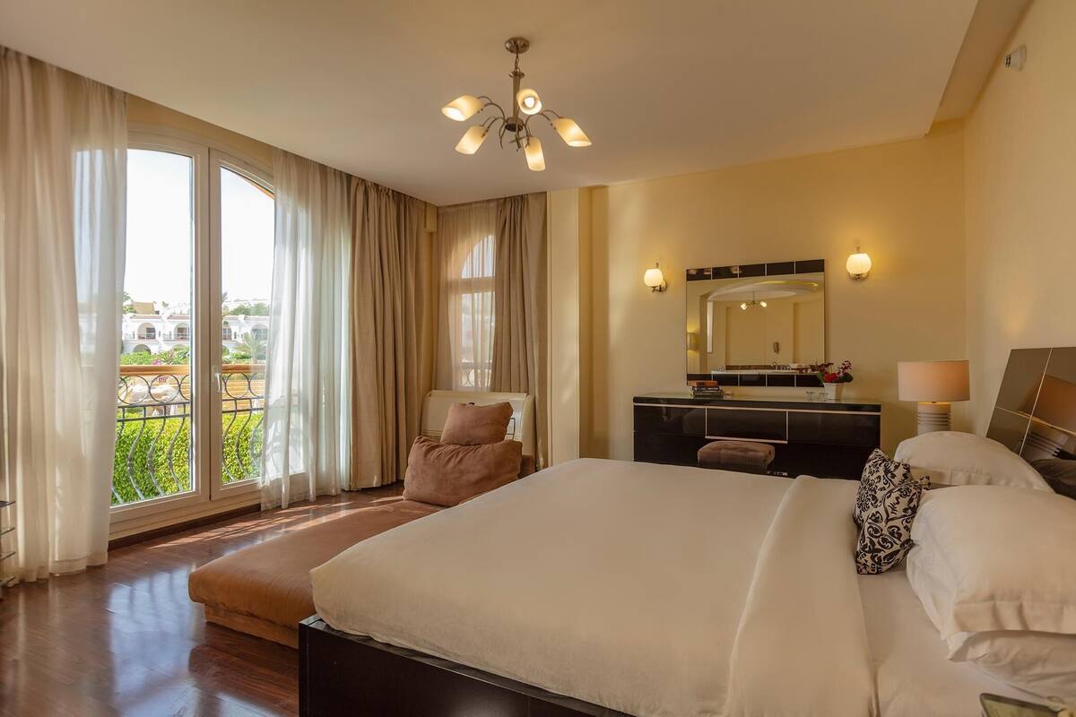 A spacious hotel room with a large bed, a sitting area, a vanity with a mirror, and a view of the outdoors.