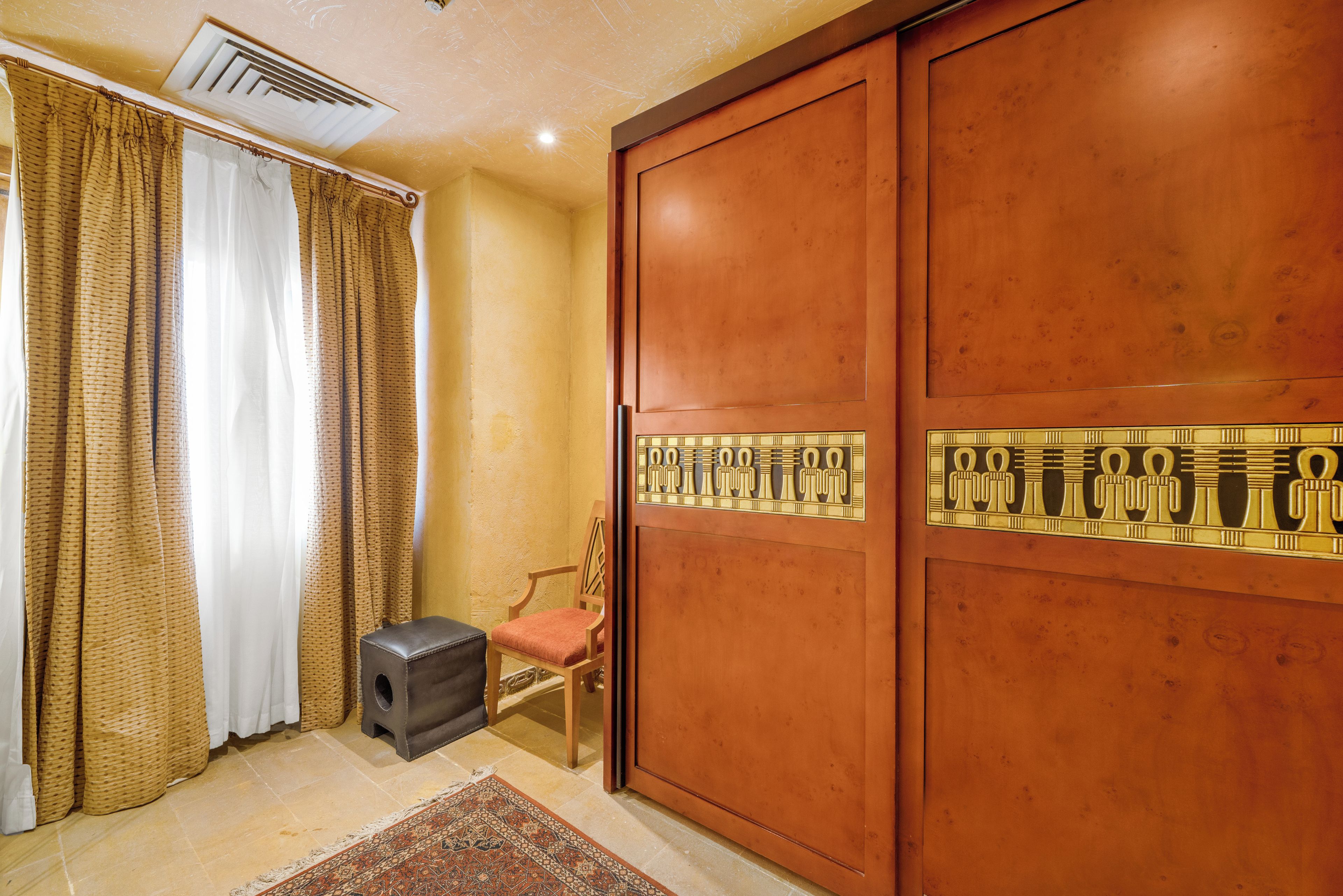 A room with a large wooden wardrobe, a small wooden chair, a black ottoman, and a patterned rug.