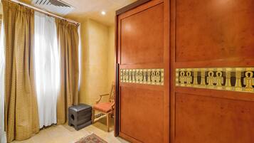 A room with a large wooden wardrobe, a small wooden chair, a black ottoman, and a patterned rug.