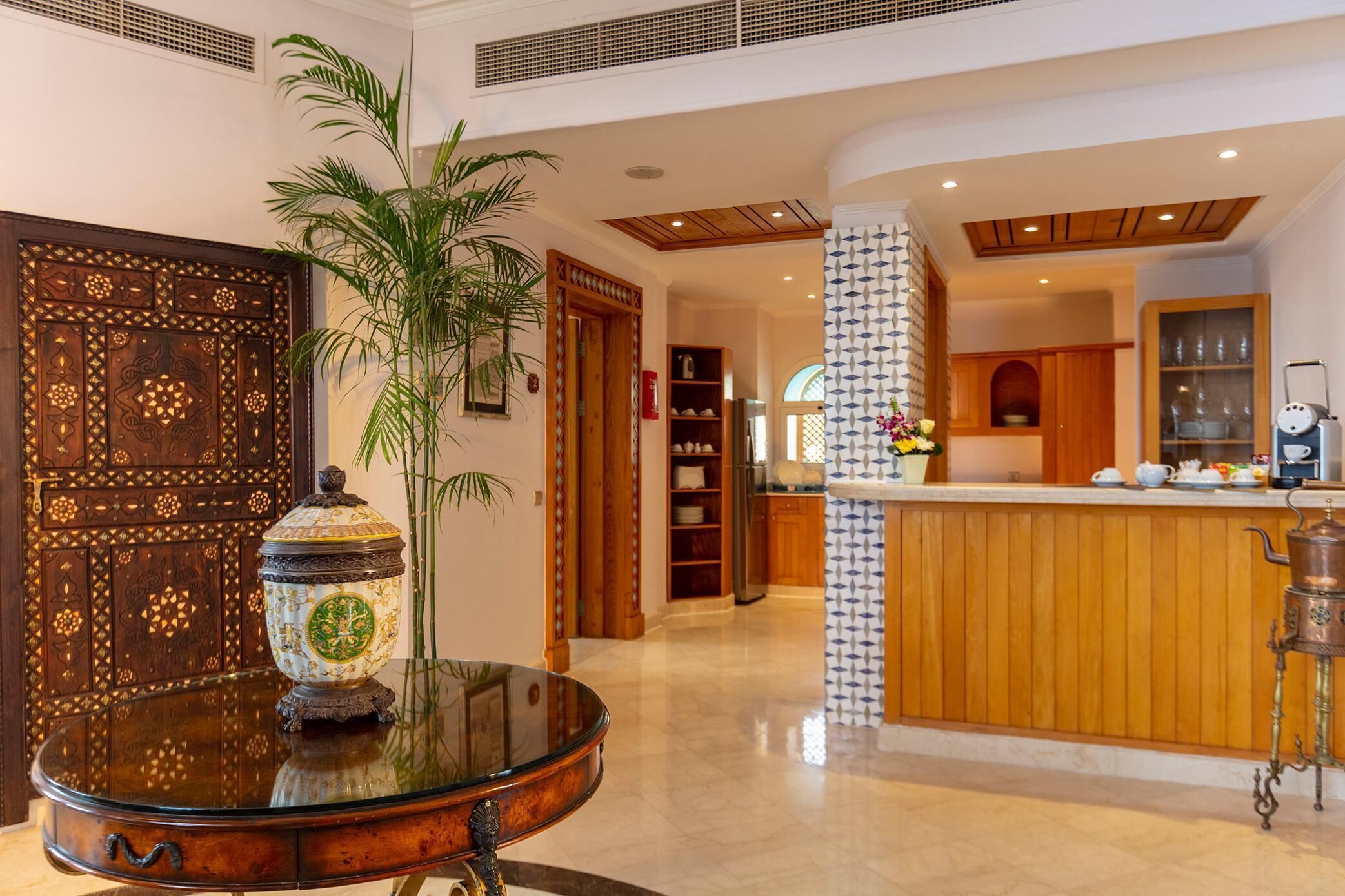 A lobby with a traditional wooden door, a glass table with a decorative item, a potted plant, and a reception area with a counter and kitchen in the background.