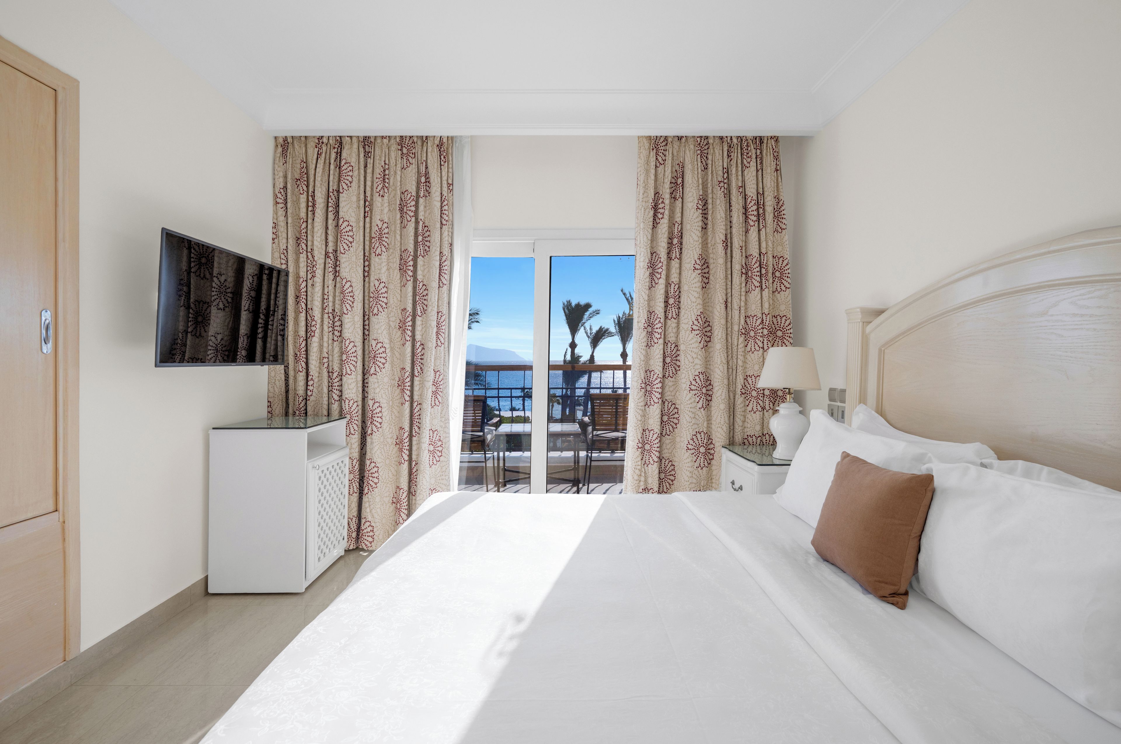 A hotel room with a bed, a TV mounted on the wall, a small desk, and a balcony with a view of palm trees and the sea.