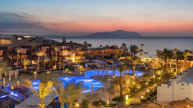 A resort with multiple swimming pools, palm trees, and a view of the sea at sunset.